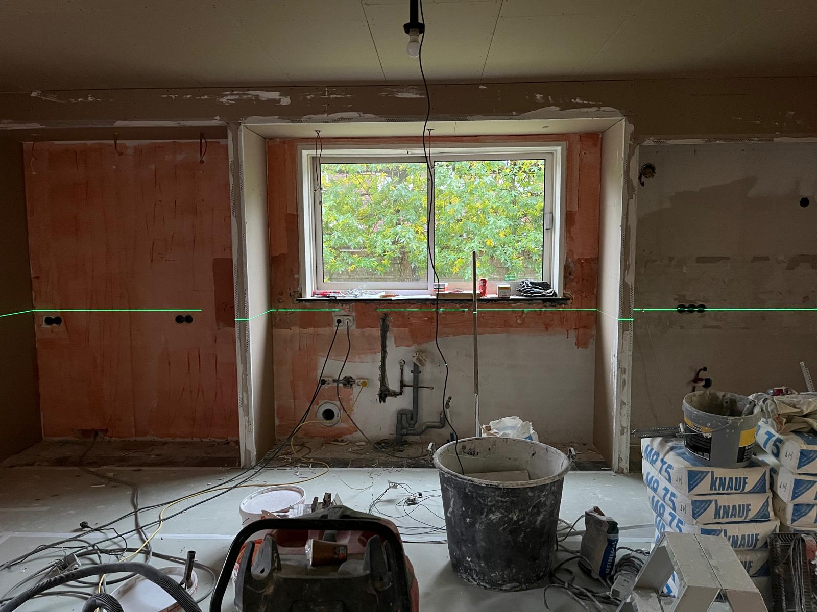 Een keukenwand tijdens het voorwerk, met een laserlijn op de wand, dozen voor stopcontacten, water en afvoer op hoogte en een omkasting met nissen rond het raam.