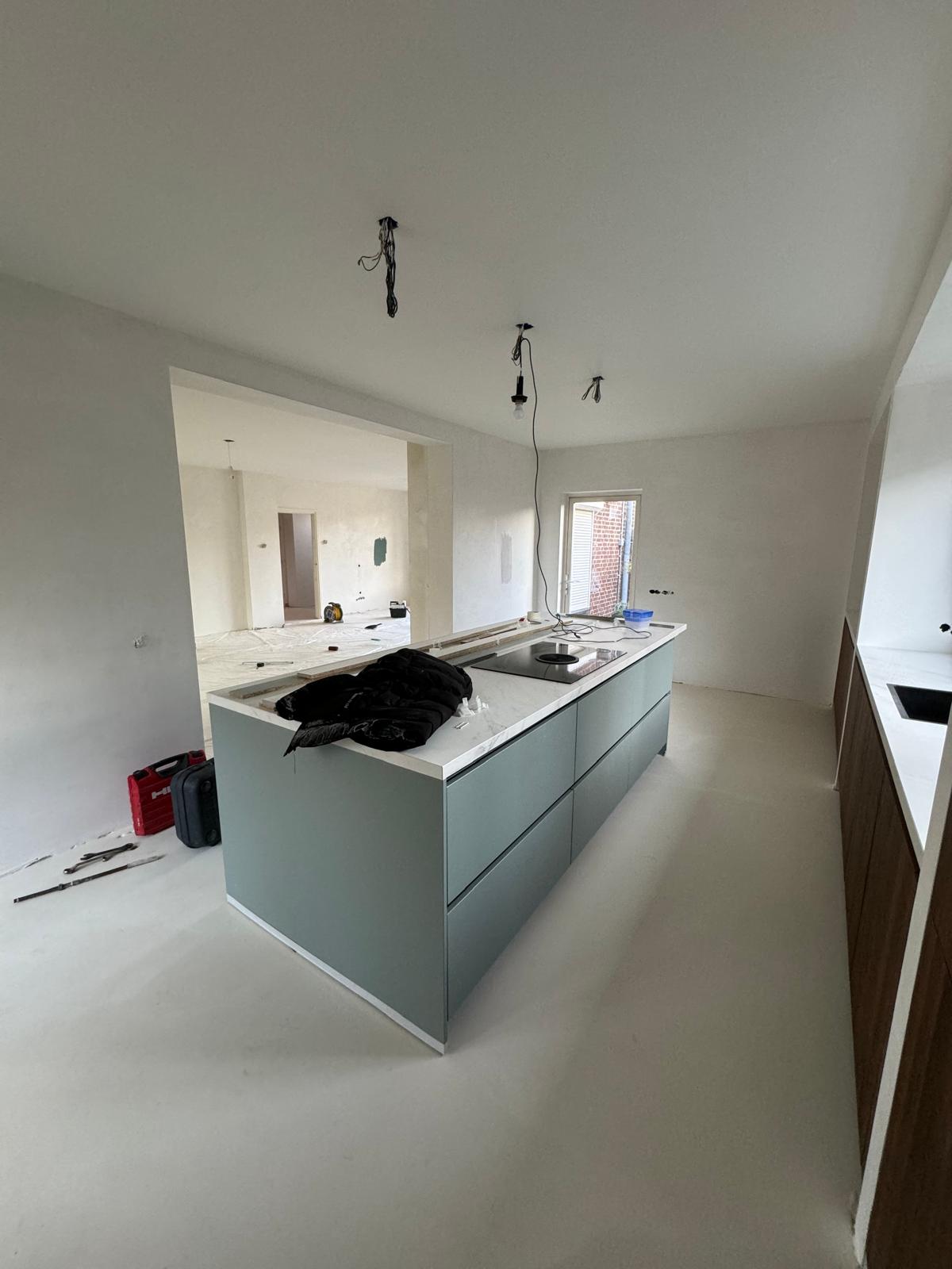 Een kookeiland met groengrijze kasten en een wit blad met een kookplaat waarin de afzuiging zit, op een lichte gietvloer. Rechts een wand met donkere houten kasten en een spoelbak, links een brede doorbraak naar de woonkamer die nog in de verbouwing is.